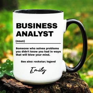 May include: White coffee mug with a black handle and black rim. The mug has a black and white graphic that reads "BUSINESS ANALYST (noun) Someone who solves problems you didn't know you had in ways that will blow your mind. See also: rockstar; legend Emily"