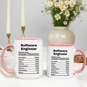 May include: Two white ceramic mugs with pink handles and rims. The mugs are printed with "Software Engineer" and a humorous "Nutrition Facts" label. A vase of white flowers sits behind the mugs.