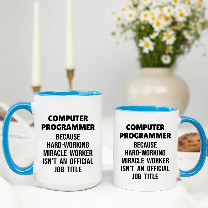 May include: Two white ceramic mugs with blue handles and rims. The mugs have the text "COMPUTER PROGRAMMER BECAUSE HARD-WORKING MIRACLE WORKER ISN'T AN OFFICIAL JOB TITLE" in black. A vase of white flowers is in the background.