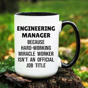 May include: White ceramic mug with a black interior and handle. The mug features the text "ENGINEERING MANAGER BECAUSE HARD-WORKING MIRACLE WORKER ISN'T AN OFFICIAL JOB TITLE" in black, bold font.