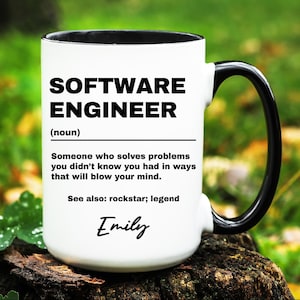 May include: White ceramic mug with a black handle and rim. The mug features the text "SOFTWARE ENGINEER" above a definition, and the name "Emily" at the bottom. The mug is sitting on a wooden surface.