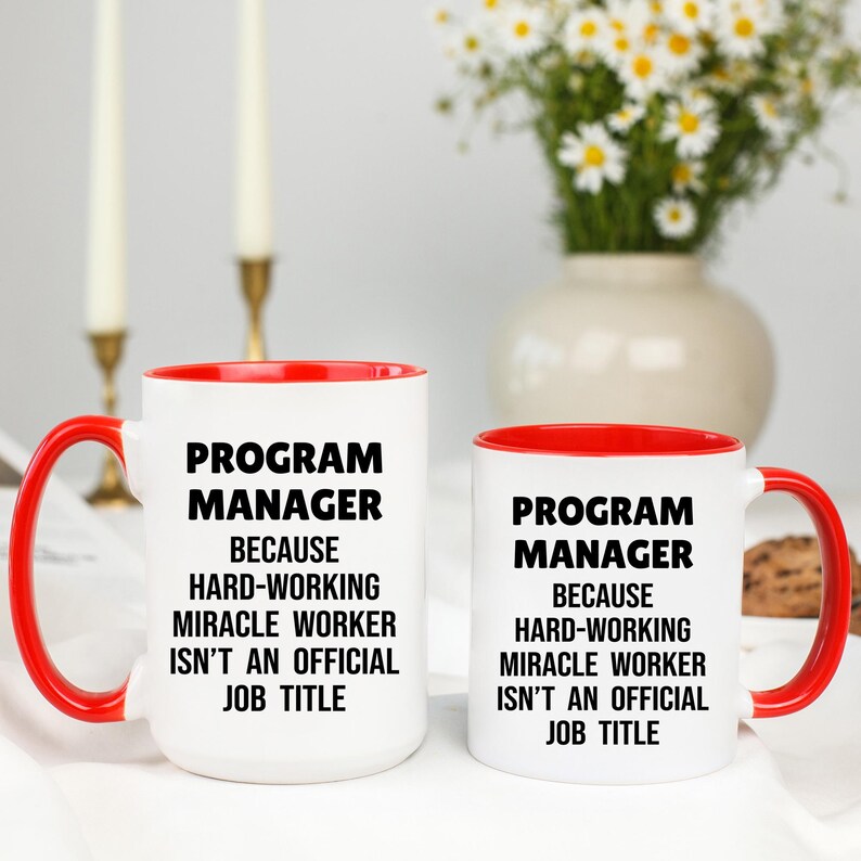 May include: Two white ceramic mugs with red interiors and handles. The mugs have the text "PROGRAM MANAGER BECAUSE HARD-WORKING MIRACLE WORKER ISN'T AN OFFICIAL JOB TITLE" in black. A vase of flowers is in the background.