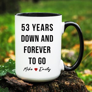 May include: A white ceramic mug with a black interior and handle. The mug features the text "53 YEARS DOWN AND FOREVER TO GO" in bold black letters. Below the text are the names "Mike" and "Emily" with a heart symbol.