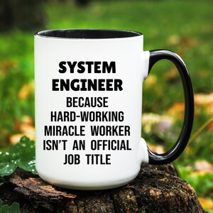 May include: White ceramic mug with a black interior and handle. The mug features the text "SYSTEM ENGINEER BECAUSE HARD-WORKING MIRACLE WORKER ISN'T AN OFFICIAL JOB TITLE" in bold black letters.