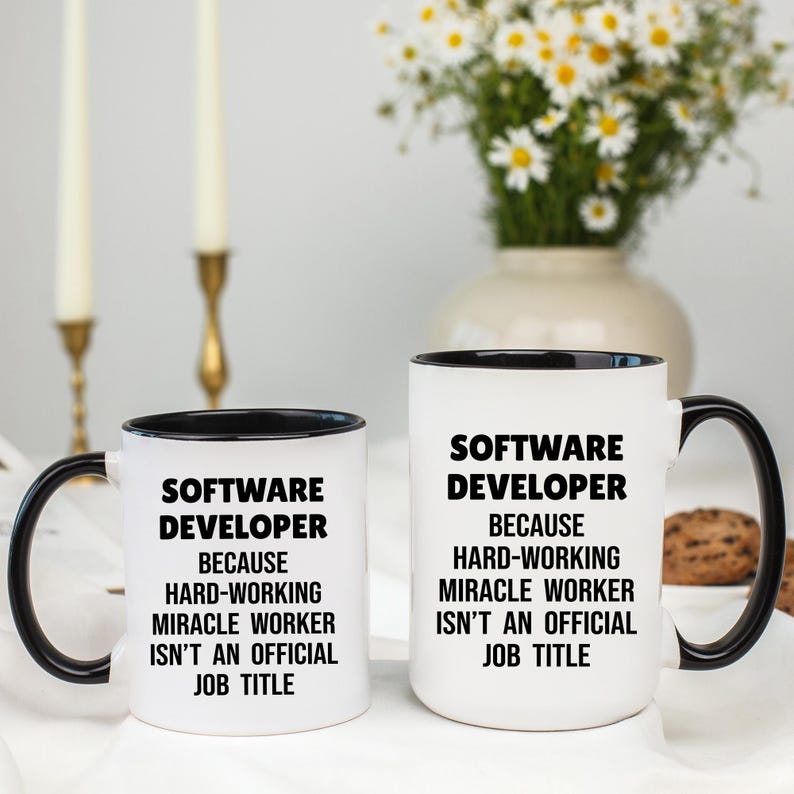 May include: Two white ceramic mugs with black handles and rims. The mugs have the text "SOFTWARE DEVELOPER BECAUSE HARD-WORKING MIRACLE WORKER ISN'T AN OFFICIAL JOB TITLE" in black. A vase of flowers and candles are in the background.