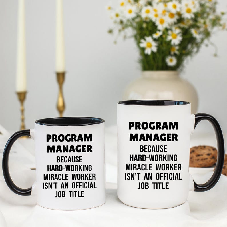May include: Two white ceramic mugs with black handles and rims. The mugs have the text "PROGRAM MANAGER" and the phrase "BECAUSE HARD-WORKING MIRACLE WORKER ISN'T AN OFFICIAL JOB TITLE" printed in black.