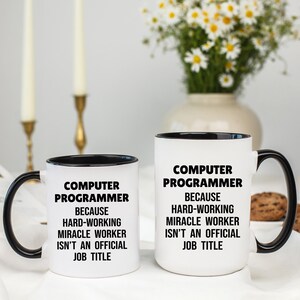May include: Two white ceramic mugs with black interiors and handles. The mugs have the text "COMPUTER PROGRAMMER BECAUSE HARD-WORKING MIRACLE WORKER ISN'T AN OFFICIAL JOB TITLE" printed in black.