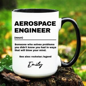 May include: White ceramic mug with a black rim and handle. The mug has black text that reads "Aerospace Engineer (noun) Someone who solves problems you didn't know you had in ways that will blow your mind. See also: rockstar; legend" and the name "Emily" written in cursive.