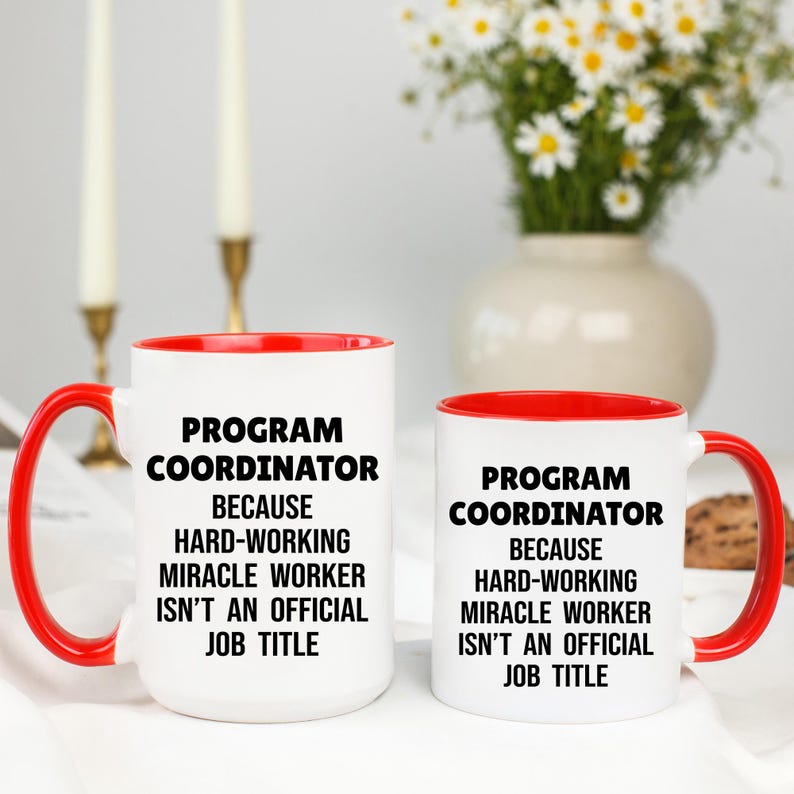 May include: Two white ceramic mugs with red interiors and handles. The mugs have the text "PROGRAM COORDINATOR BECAUSE HARD-WORKING MIRACLE WORKER ISN'T AN OFFICIAL JOB TITLE" in black. A vase of flowers is in the background.