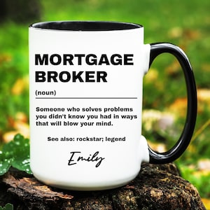 May include: A white ceramic mug with a black handle and rim. The mug features the words "MORTGAGE BROKER" in bold black letters, along with a definition and the name "Emily" in script. The mug is sitting on a wooden surface.
