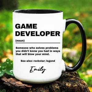 May include: White ceramic mug with a black rim and handle. The mug has a black graphic that reads "GAME DEVELOPER (noun) Someone who solves problems you didn't know you had in ways that will blow your mind. See also: rockstar; legend" with the name "Emily" written in cursive below the text.