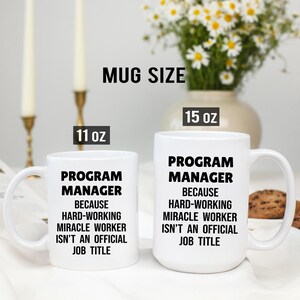 May include: Two white ceramic mugs with black text that reads "PROGRAM MANAGER BECAUSE HARD-WORKING MIRACLE WORKER ISN'T AN OFFICIAL JOB TITLE." One mug is 11 oz, the other is 15 oz. A vase of flowers is in the background.
