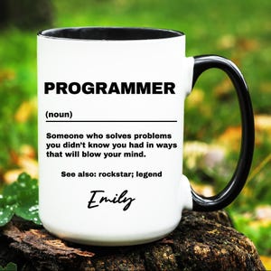 May include: White ceramic mug with a black interior and handle. The mug features the word "PROGRAMMER" in bold black letters, along with a definition and the name "Emily" in cursive. The mug is set against a blurred green background.