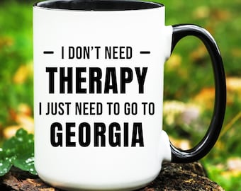 Może przedstawiać: Biały ceramiczny kubek z czarnym wnętrzem i uchwytem. Kubek zawiera tekst "I DON'T NEED THERAPY I JUST NEED TO GO TO GEORGIA" pogrubionymi czarnymi literami. Kubek stoi na drewnianej powierzchni.