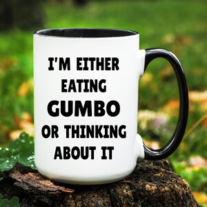 May include: White ceramic mug with a black interior and handle. The mug features the text "I'M EITHER EATING GUMBO OR THINKING ABOUT IT" in bold black letters. The mug is sitting on a piece of wood, with a blurred green background.