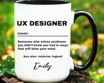 May include: White ceramic mug with a black interior and handle. The mug features the text "UX DESIGNER" above a definition, and the name "Emily" in script. The mug is sitting on a wooden surface.