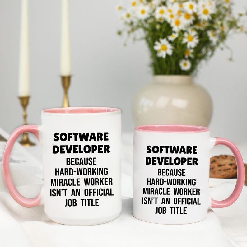 May include: Two white coffee mugs with pink interiors and handles. The mugs feature the text "SOFTWARE DEVELOPER BECAUSE HARD-WORKING MIRACLE WORKER ISN'T AN OFFICIAL JOB TITLE" in black. A vase of white flowers is in the background.