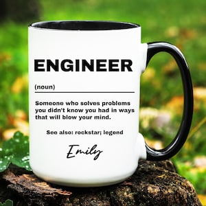 May include: White ceramic mug with a black interior and handle. The mug features the word "ENGINEER" in bold black letters, along with a definition and the name "Emily" in script. The mug is sitting on a wooden surface.