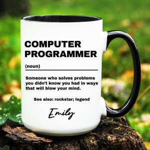 May include: White ceramic mug with a black handle and rim. The mug features the text "COMPUTER PROGRAMMER (noun)" along with a humorous definition and the name "Emily". The mug is sitting on a wooden surface.