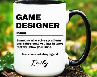 May include: White ceramic mug with a black rim and handle. The mug has black text that reads "GAME DESIGNER (noun) Someone who solves problems you didn't know you had in ways that will blow your mind. See also: rockstar; legend Emily"