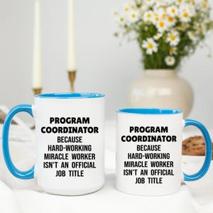 May include: Two white ceramic mugs with blue handles and rims. The mugs have the text "PROGRAM COORDINATOR BECAUSE HARD-WORKING MIRACLE WORKER ISN'T AN OFFICIAL JOB TITLE" in black. A vase of white flowers is in the background.