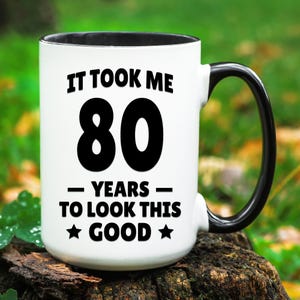 May include: White ceramic mug with a black handle and black rim. The mug has black text that reads "It took me 80 years to look this good." with a star on either side of the word "good".
