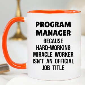 May include: White ceramic mug with an orange handle and rim. The mug features the text "PROGRAM MANAGER" above the phrase "BECAUSE HARD-WORKING MIRACLE WORKER ISN'T AN OFFICIAL JOB TITLE" in black, bold font.