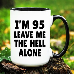 May include: White ceramic mug with a black interior and handle. The mug features the text "I'M 95 LEAVE ME THE HELL ALONE" in bold black letters. The mug is sitting on a wooden surface against a blurred green background.