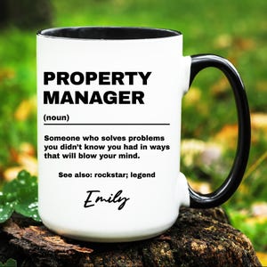 May include: White ceramic mug with a black handle and rim. The mug features the text "PROPERTY MANAGER" in bold black letters, along with a definition and the name "Emily" in script. The mug is sitting on a wooden surface.