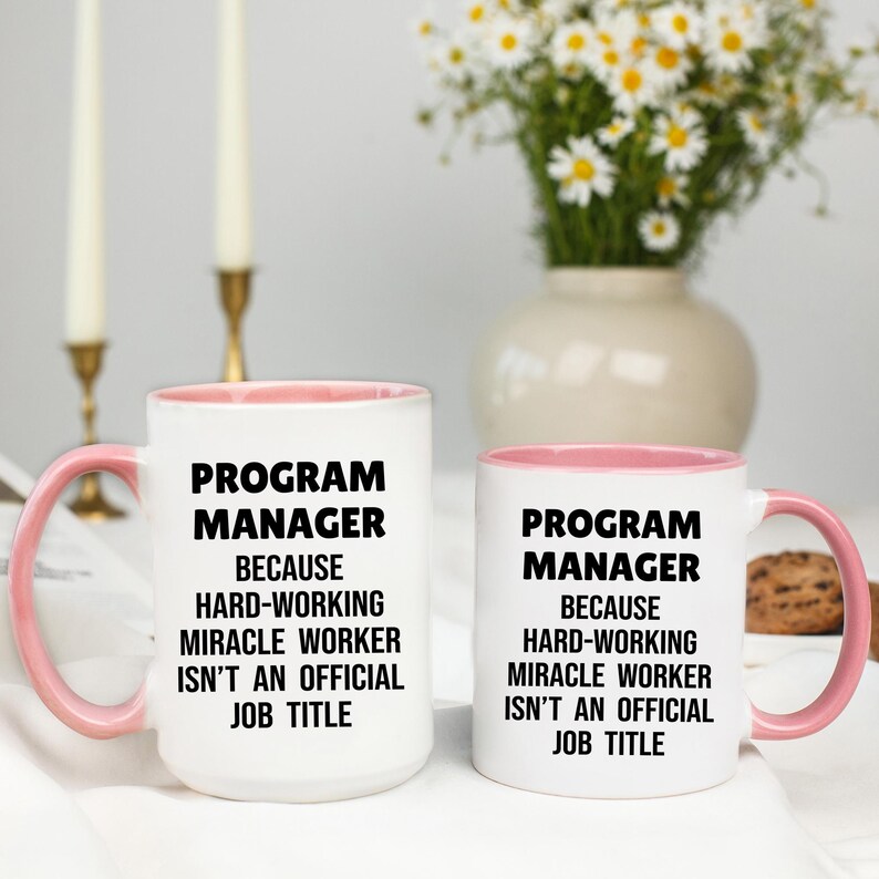 May include: Two white ceramic mugs with pink interiors and handles. The mugs have the text "PROGRAM MANAGER BECAUSE HARD-WORKING MIRACLE WORKER ISN'T AN OFFICIAL JOB TITLE" in black. A vase of white flowers is in the background.