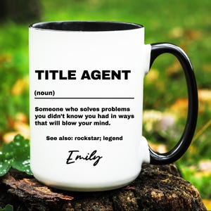 May include: White ceramic mug with a black interior and handle. The mug features the text "TITLE AGENT" above a definition, and the name "Emily" in cursive. The mug is sitting on a wooden surface.