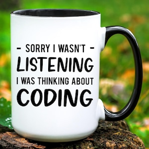 May include: White ceramic mug with a black handle and black rim. The mug has black text that reads "Sorry I wasn't listening I was thinking about coding."