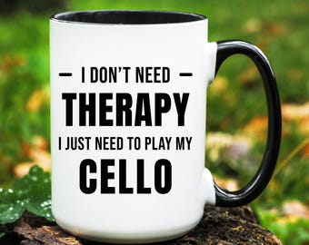 May include: White ceramic mug with a black interior and handle. The mug features the text "I DON'T NEED THERAPY I JUST NEED TO PLAY MY CELLO" in bold black letters. The mug is sitting on a wooden surface with a green background.