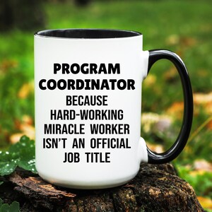 May include: White ceramic mug with a black interior and handle. The mug features the text "PROGRAM COORDINATOR BECAUSE HARD-WORKING MIRACLE WORKER ISN'T AN OFFICIAL JOB TITLE" in black, centered on the front. The mug is set against a blurred green background.