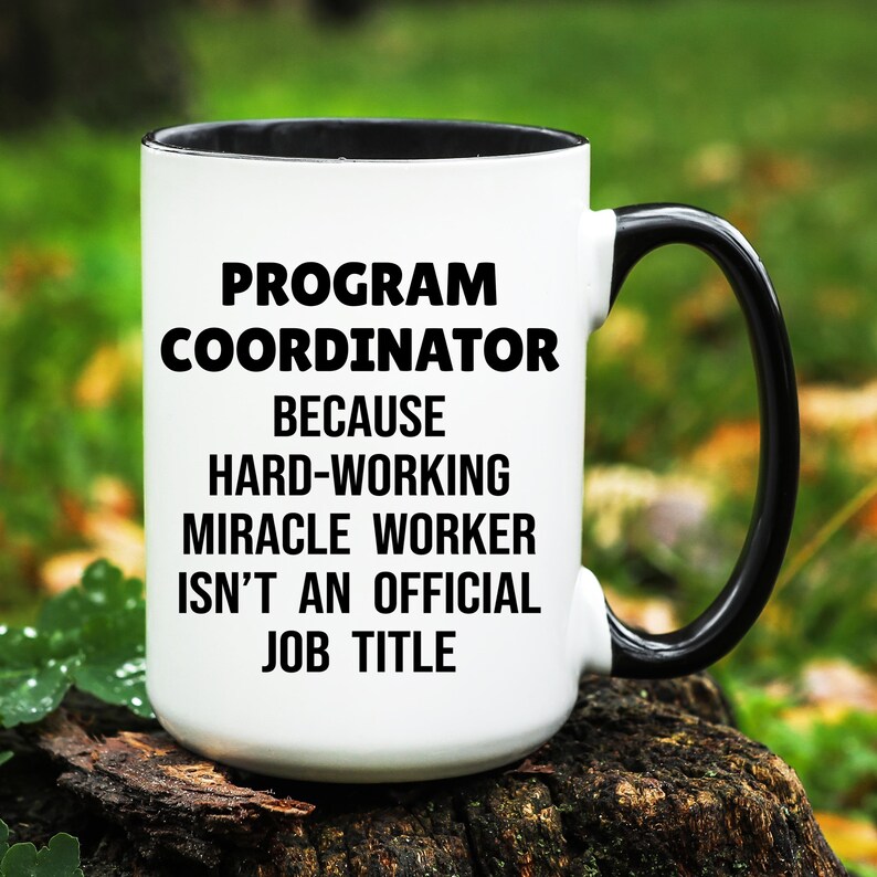 May include: White ceramic mug with a black interior and handle. The mug features the text "PROGRAM COORDINATOR BECAUSE HARD-WORKING MIRACLE WORKER ISN'T AN OFFICIAL JOB TITLE" in black, centered on the front. The mug is set against a blurred green background.