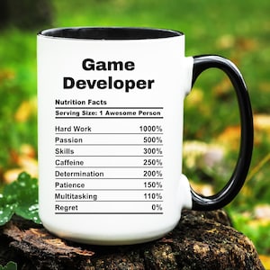 May include: White ceramic mug with a black handle. The mug has a black rim and features a nutrition facts label for "Game Developer" with a serving size of "1 Awesome Person". The label lists the percentage of ingredients such as hard work, passion, skills, caffeine, determination, patience, multitasking, and regret.