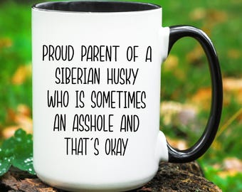 May include: White ceramic mug with a black handle and rim. The mug features the text "PROUD PARENT OF A SIBERIAN HUSKY WHO IS SOMETIMES AN ASSHOLE AND THAT'S OKAY" in black lettering. The mug is sitting on a wooden surface.