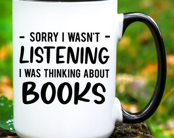 May include: White ceramic coffee mug with a black rim and handle. The mug has black text that reads "Sorry I wasn't listening I was thinking about books".