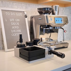 May include: A silver espresso machine with a digital screen sits next to a grey felt letter board displaying the text TO DO LIST 1. SIT ON DECK AND DRINK COFFEE. In front is a light grey and black coffee station organiser holding a portafilter.