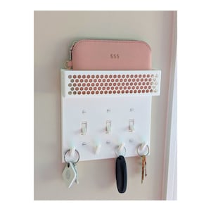 Könnte beinhalten: Weißer, an der Wand befestigter Schlüsselhalter mit einem Regal und drei Haken im Lichtschalter-Stil. Eine rosa Geldbörse mit "$$$" liegt auf dem Regal. Schlüssel und ein Autoschlüssel hängen an den Haken. Das Regal hat ein Wabenmuster.