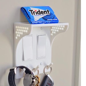 Könnte beinhalten: Weiße Wandhalterung aus Kunststoff mit zwei Lichtschaltern, einem Regal für einen kleinen Gegenstand und drei Haken zum Aufhängen von Schlüsseln oder anderen kleinen Gegenständen. Eine Packung Trident Original Flavor Kaugummi befindet sich auf dem Regal. Eine Uhr und Schlüssel hängen an den Haken.