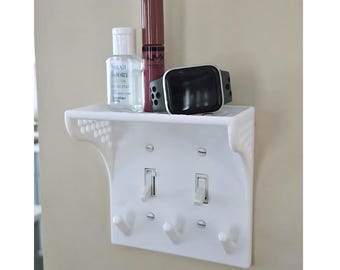 Puede incluir: Estante de plástico blanco con ganchos y dos interruptores de luz. El estante está montado sobre una placa de interruptor de luz. Hay tres artículos en el estante: una pequeña botella de desinfectante para manos, un tubo de lápiz labial y un reloj inteligente.