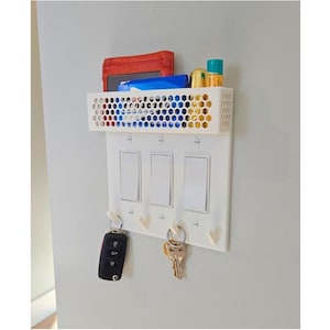 Könnte beinhalten: Weißer, wandmontierter Organizer aus Kunststoff mit einem Drahtkorb, drei Lichtschaltern und vier Haken für Schlüssel. Der Korb enthält eine rote Brieftasche, ein blaues Paket und zwei kleine Flaschen.