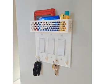 Puede incluir: Organizador de pared blanco de plástico con una cesta de alambre, tres tapas de interruptor de luz y cuatro ganchos para llaves. La cesta contiene una cartera roja, un paquete azul y dos botellas pequeñas.