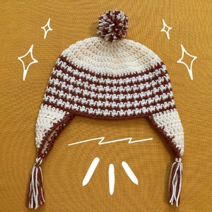 May include: A brown and white striped crocheted hat with a pom pom on top and ear flaps. The hat has tassels on the ends of the ear flaps.