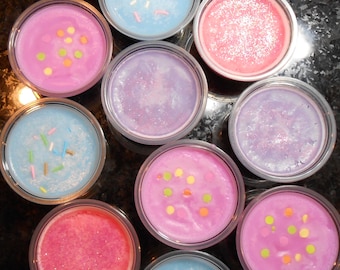May include: Ten small round containers of wax melts, each with a different color and design. The colors include pink, blue, purple, and yellow. Some of the wax melts have sprinkles on top, while others have glitter.