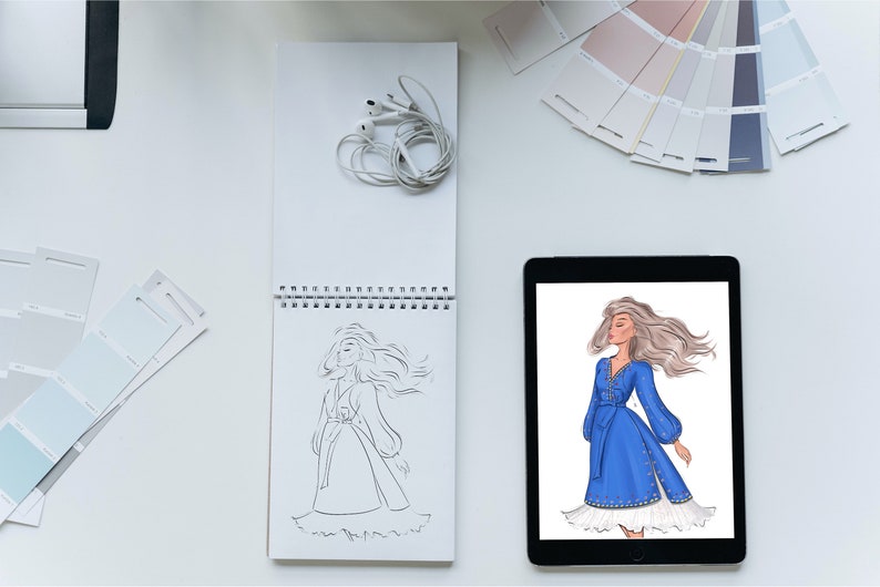 May include: A digital tablet displaying a fashion illustration of a woman wearing a blue dress with white embroidery. The woman has long blonde hair and is standing with her arms at her sides. The tablet is on a white surface next to a sketchbook with a pencil drawing of a woman in a dress.