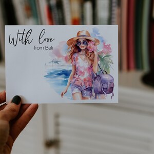 May include: A watercolour illustration of a woman wearing a straw hat, sunglasses, and a pink floral shirt. She is carrying a purple backpack and walking on a beach with palm trees in the background. The text "With love from Bali" is written on the card.