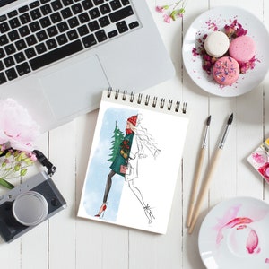May include: A sketchbook with a drawing of a woman in a winter outfit holding a Christmas tree. The sketchbook is on a white table with a laptop, a camera, flowers, a cup of tea, macarons, paintbrushes, and watercolor paints.
