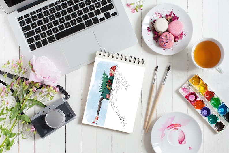 May include: A sketchbook with a drawing of a woman in a winter outfit holding a Christmas tree. The sketchbook is on a white table with a laptop, a camera, flowers, a cup of tea, macarons, paintbrushes, and watercolor paints.
